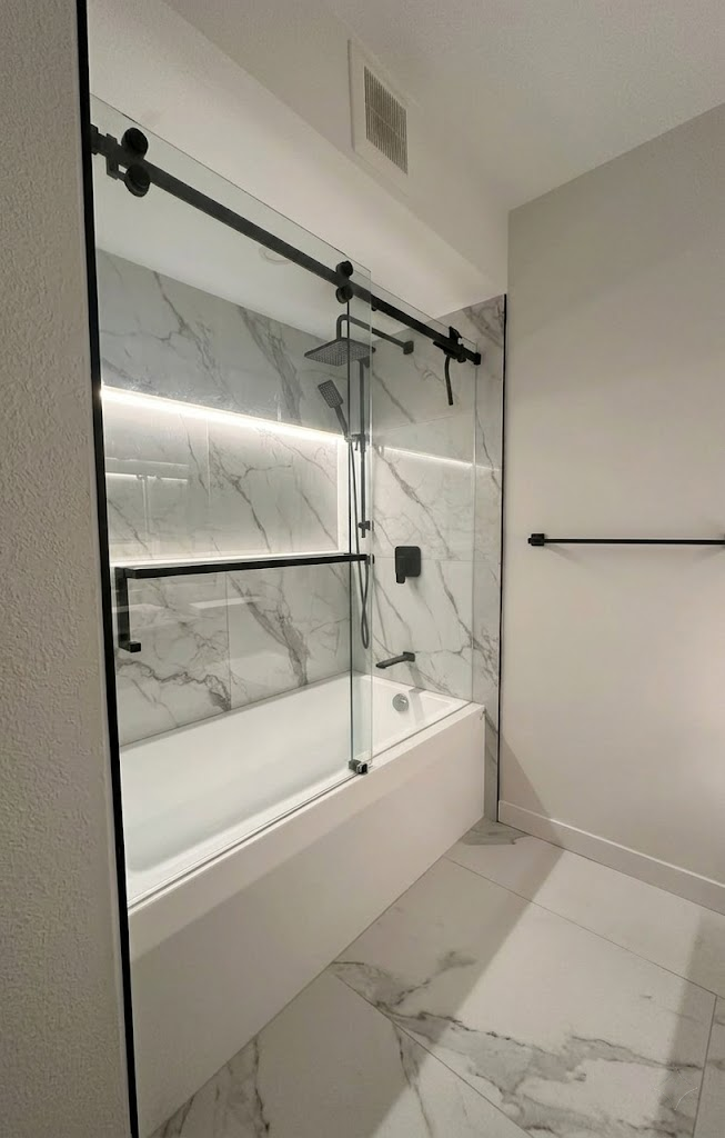 Rebuilt bathroom with a new tub, marble-look surround, lit niche, and matte black fixtures behind a sliding glass door
